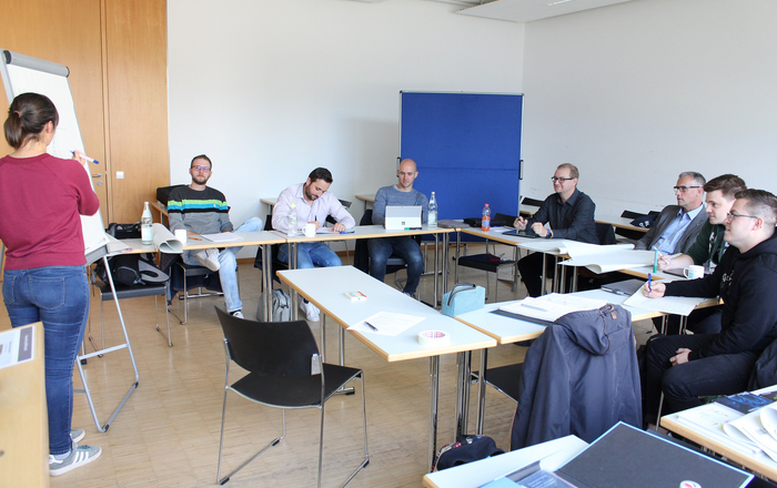 Eine Studierende steht an einer Flipchart, die anderen Studierenden sitzen an Tischen