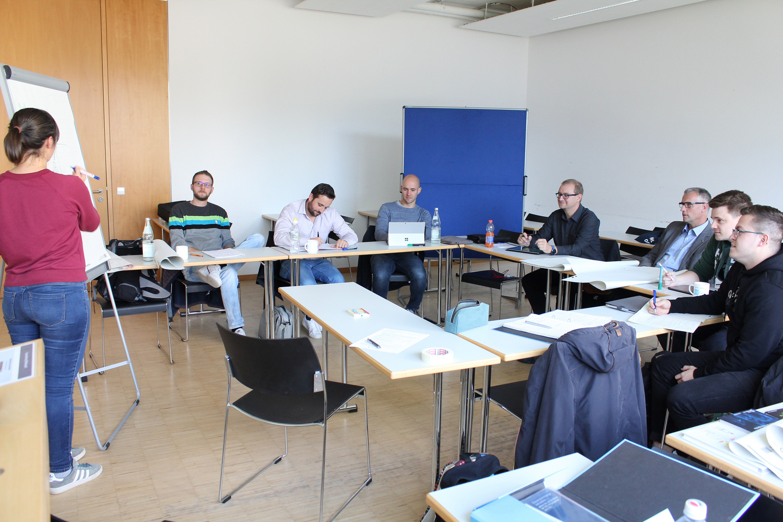 Eine Studierende steht an einer Flipchart, die anderen Studierenden sitzen an Tischen