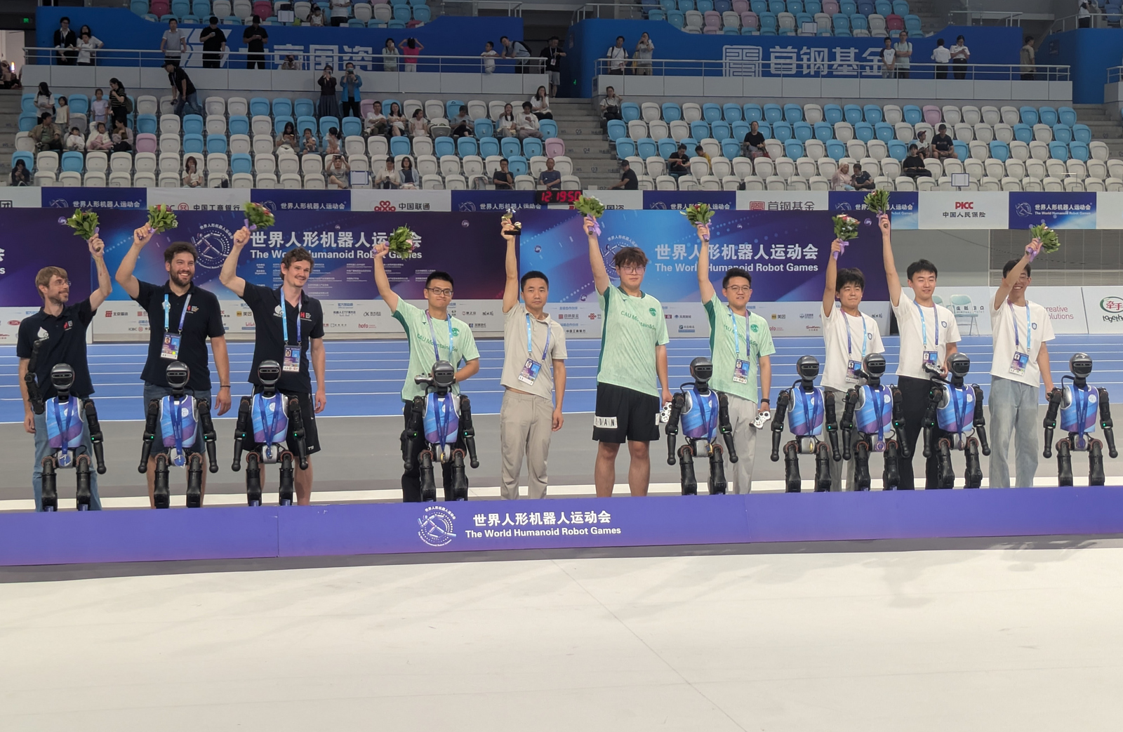 Neun junge Männer stehen hinter neun humanoiden Robotern und recken jubelnd kleine Blumensträuße in die Luft