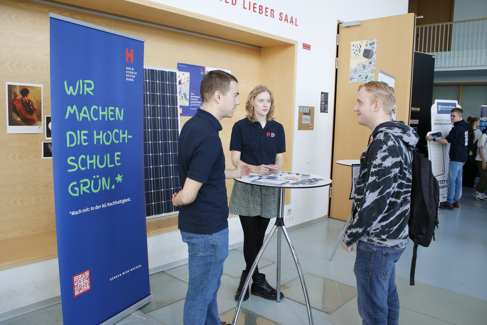 Ein Mann und eine junge Frau erklären einem blonden jungen Mann am Infotisch der AG Nachhaltigkeit, was diese macht