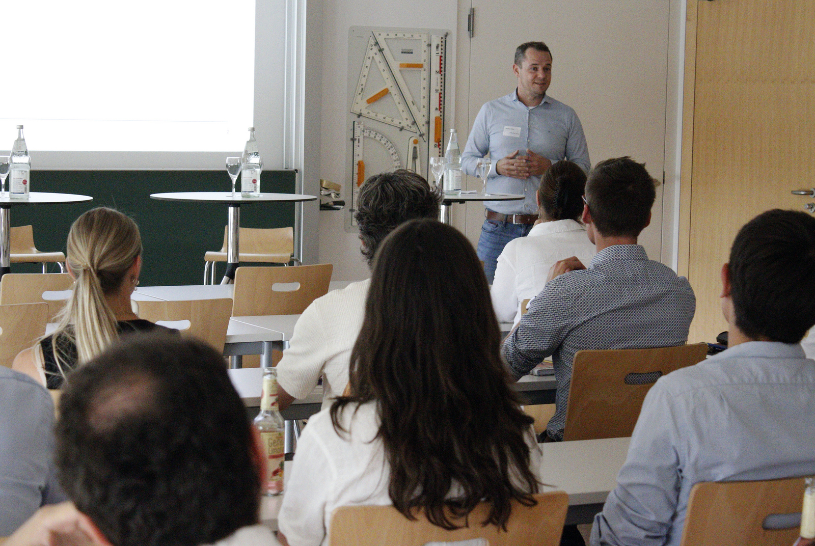 Ein Mann steht in einem Hörsaal vor einer Gruppe sitzender Studierender und spricht zu ihnen