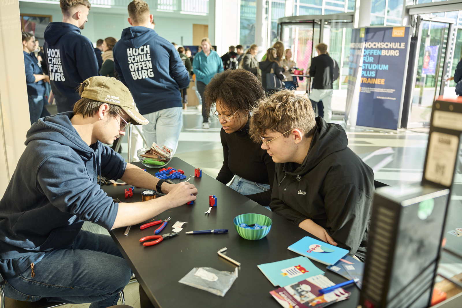 Ein Student, eine Schülerin und ein Schüler sitzen an einem Tisch und bauen einen kleinen Elektromotor