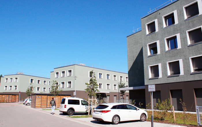 Drei grün-braune Gebäude, davor Parkplätze und zwei Fahrradunterstände aus Holz 