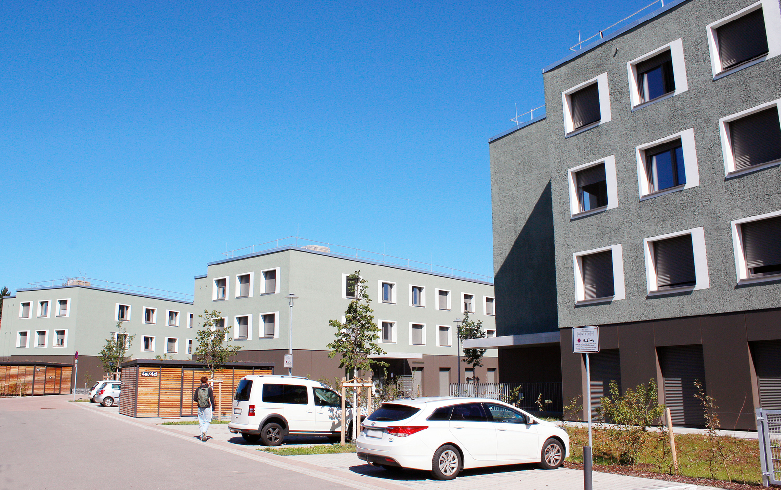 Drei grün-braune Gebäude, davor Parkplätze und zwei Fahrradunterstände aus Holz 