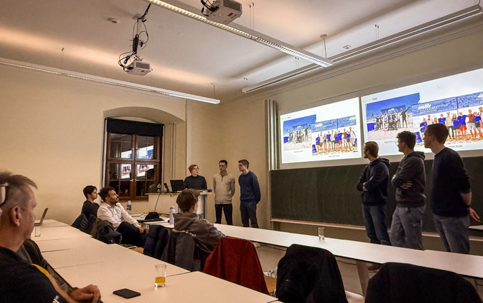 Studierende stehen neben eine Leinwand und erklären sitzenden Zuhörenden eine Präsentation