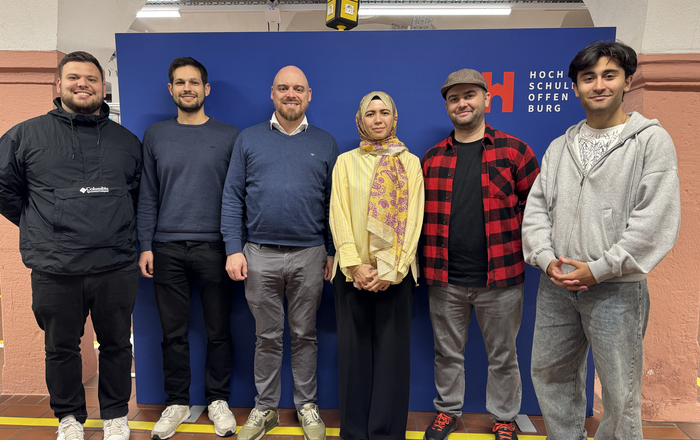 Sechs Personen stehen nebeneinander vor einer blauen Wand mit dem Schriftzug 'Hochschule Offenburg'.