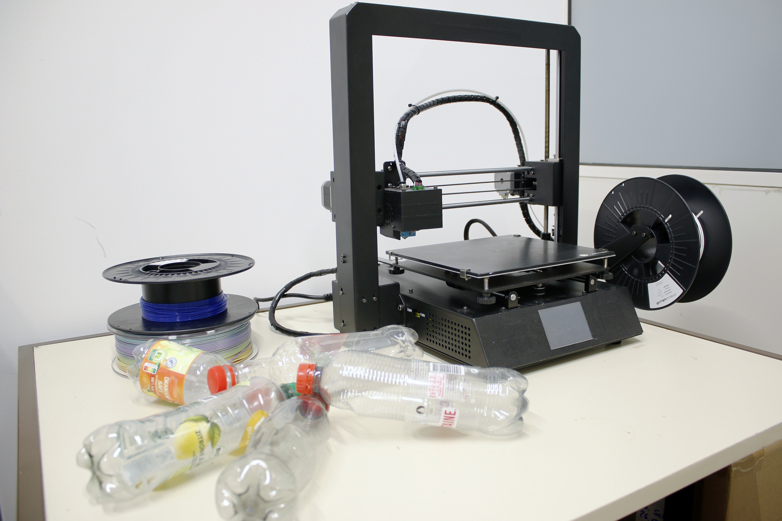 Links neben einem 3D-Drucker liegen einige alte Plastikflaschen und dahinter zwei Rollen Filamente.