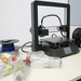 Links neben einem 3D-Drucker liegen einige alte Plastikflaschen und dahinter zwei Rollen Filamente.