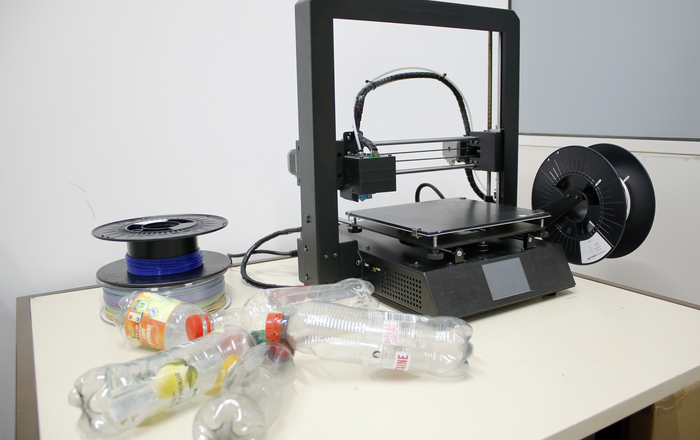 Links neben einem 3D-Drucker liegen einige alte Plastikflaschen und dahinter zwei Rollen Filamente.
