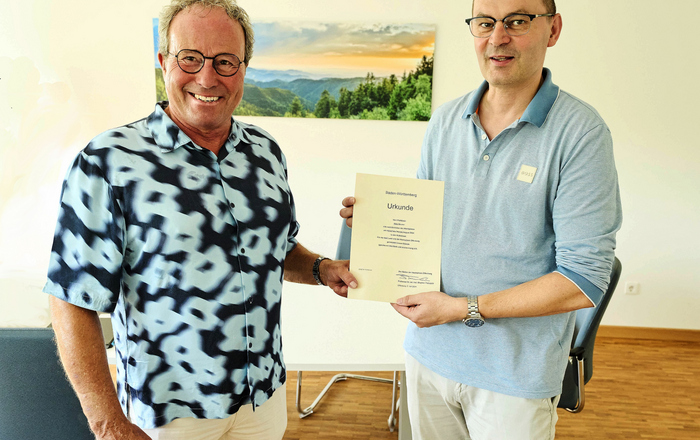 Zwei Männer stehen vor einem Schwarzwaldfoto und halten eine Urkunde zwischen sich