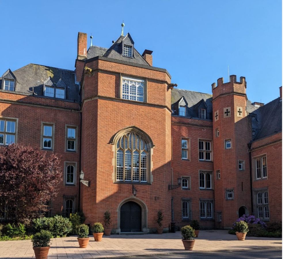 Ein rotes Backsteingebäude mit einem gewölbten Eingang und mehreren Fenstern unter einem klaren blauen Himmel, umgeben von Topfpflanzen und einer Grasfläche im Vordergrund.