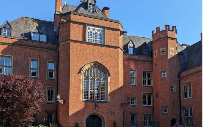 Ein rotes Backsteingebäude mit einem gewölbten Eingang und mehreren Fenstern unter einem klaren blauen Himmel, umgeben von Topfpflanzen und einer Grasfläche im Vordergrund.