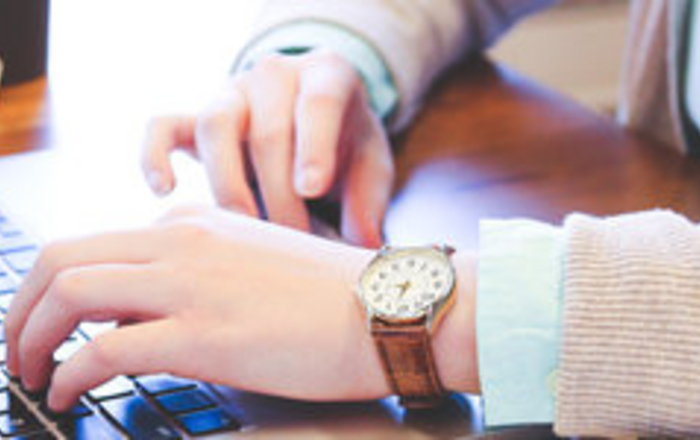 Jemand, von dem nur die Unterarme zu sehen sind, sitzt an einem Tisch und tippt auf einem Laptop