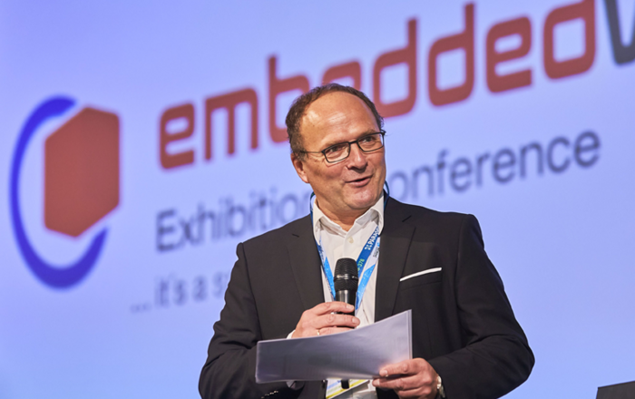 Prof. Sikora mit Mikrofon vor "embeddedworld" Hintergund
