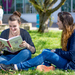 Zwei Studentinnen sitzen auf der Campus-Wiese, eine liest in einem Buch.