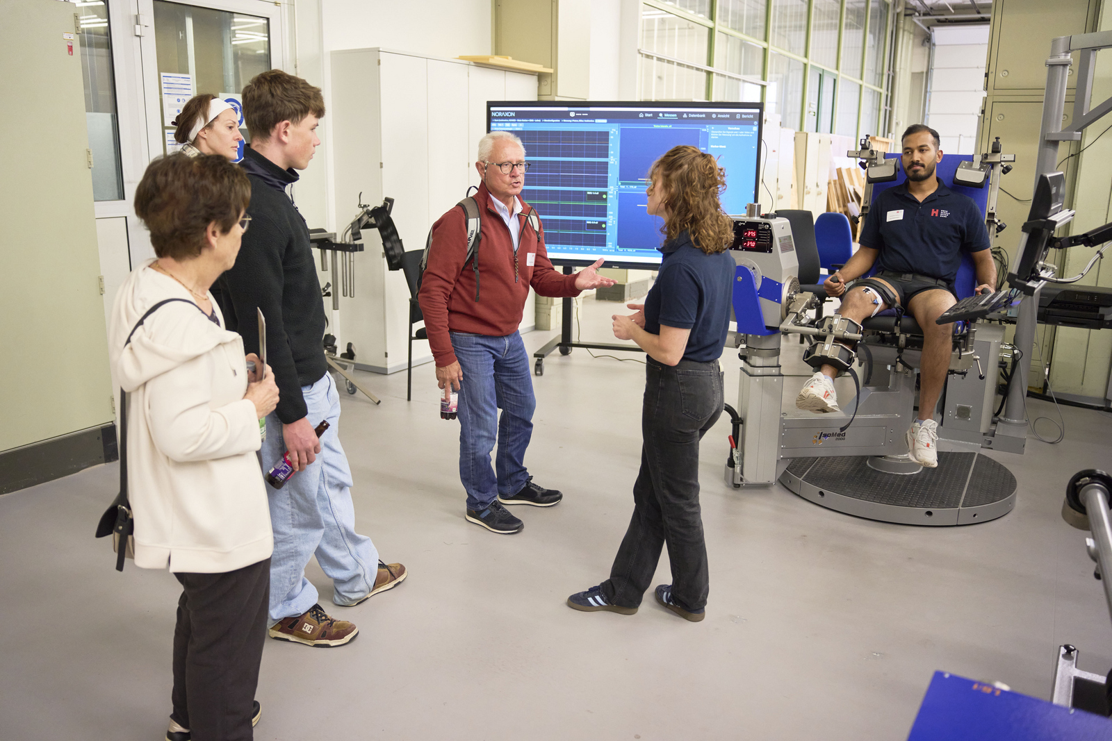 Menschen stehen neben einem Behandlungsstuhl und einem Bildschirm, eine Frau erklärt etwas