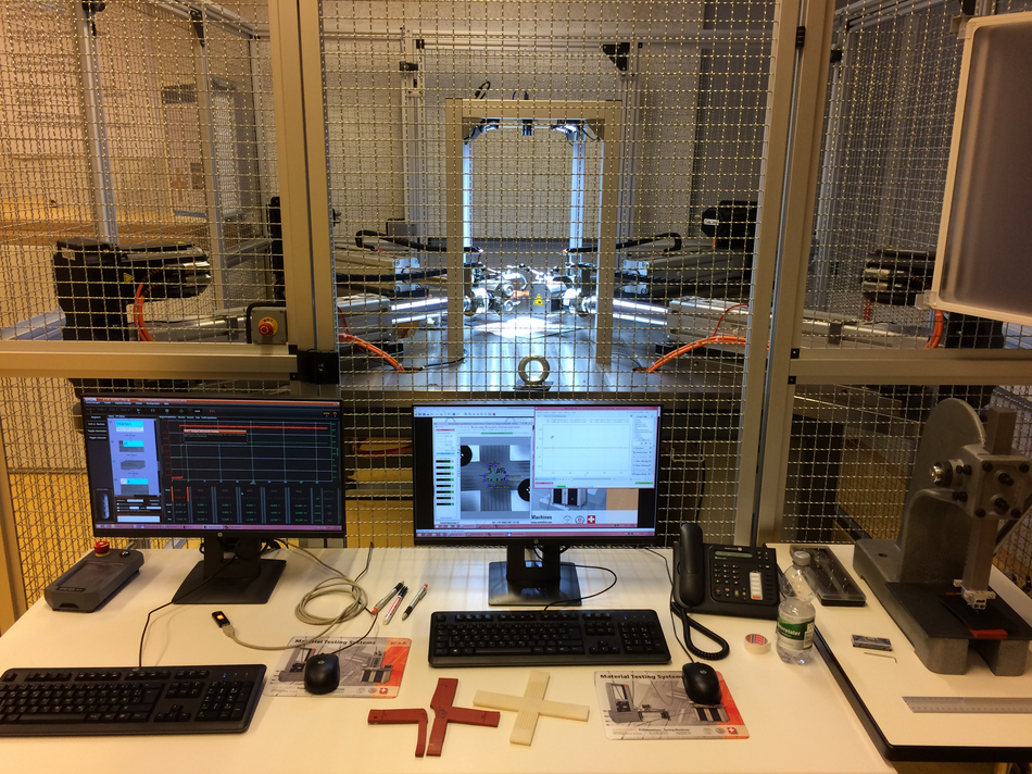 Tisch mit Computerbildschirmen und -tastaturen dahinter ein Metallkäfig in dem die zweiachsige Prüfmaschine LFM-BIAX steht.