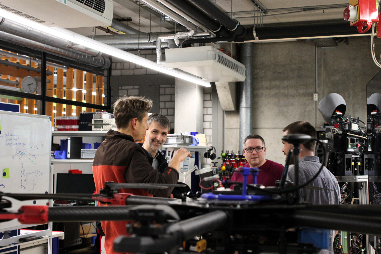 Vier junge Männer unterhalten sich in einem Labor in dem zwei humanoide Roboter stehen 