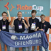 Vier Männer stehen mit Teamfahne und Pokal vor einer RoboCup Wand und jubeln