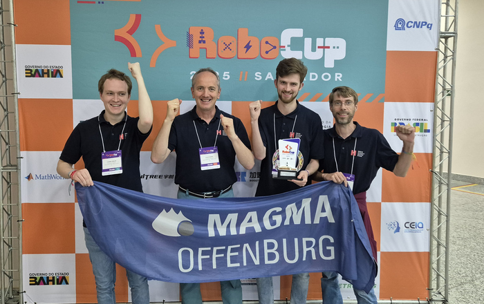 Vier Männer stehen mit Teamfahne und Pokal vor einer RoboCup Wand und jubeln