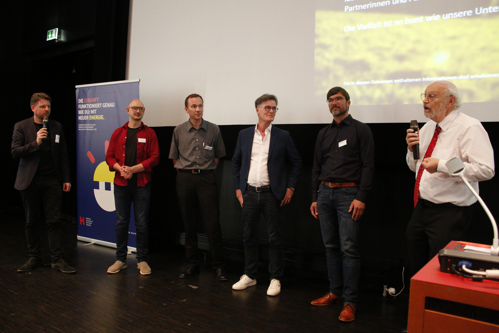 Der Moderator und fünf der sechs Vortragenden stehen in einer Reihe vor der Leinwand.