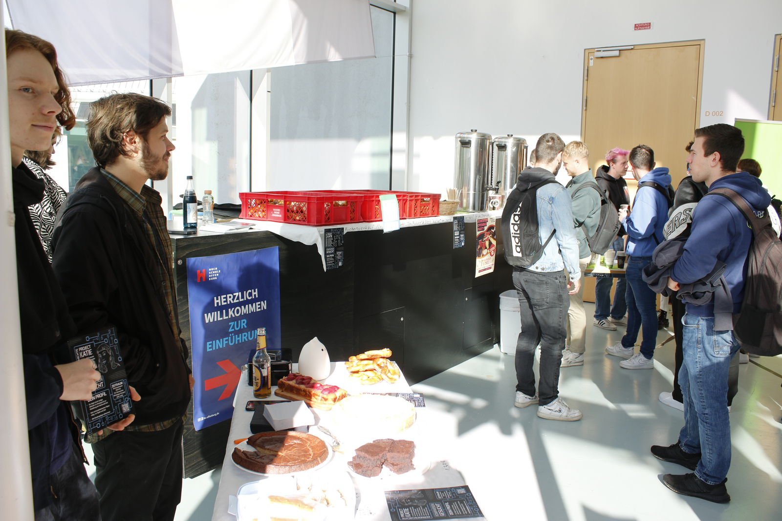 Studierende stehen hinter einem Tisch mit Kuchen, im Hintergund stehen auf einem Tresen große Kaffeekannen