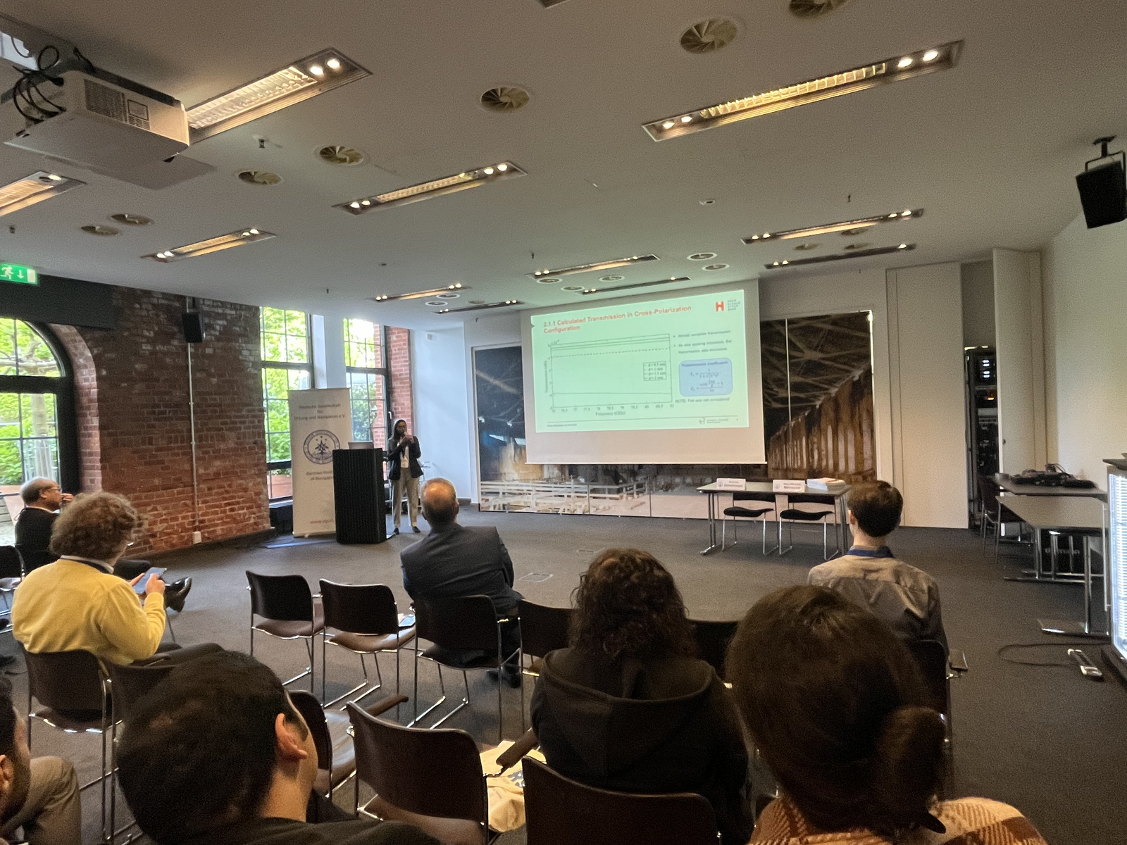 Eine Frau steht vor einer Leinwand neben einem Rednerpult und hält einen Vortrag vor sitzenden Zuhörenden