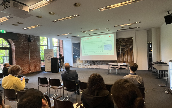 Eine Frau steht vor einer Leinwand neben einem Rednerpult und hält einen Vortrag vor sitzenden Zuhörenden