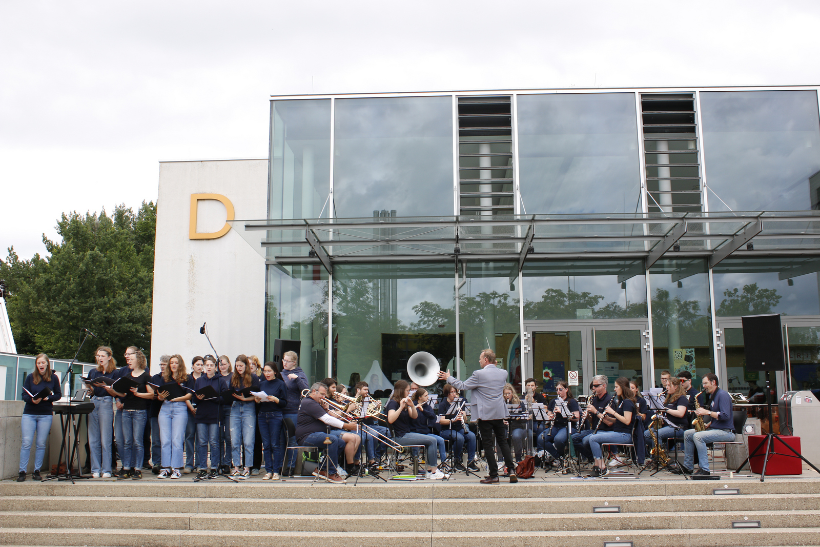 Chor und Orchester stehen beziehungsweise sitzen auf der Treppe vor Gebäude D.