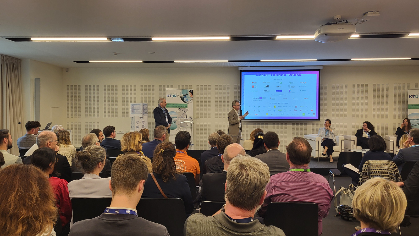 Zwei Herren stehen vor einer Leinwand vor sitzendem Publikum