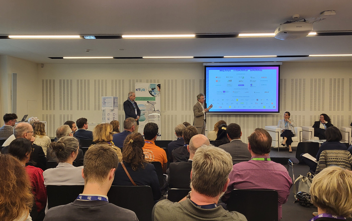 Zwei Herren stehen vor einer Leinwand vor sitzendem Publikum