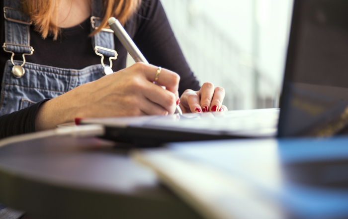 Das Bild zeigt den Oberkörper und die Hände einer an einem Tisch vor einem Laptop sitzenden Frau in Jeans-Latzhose, die etwas aufschreibt. 