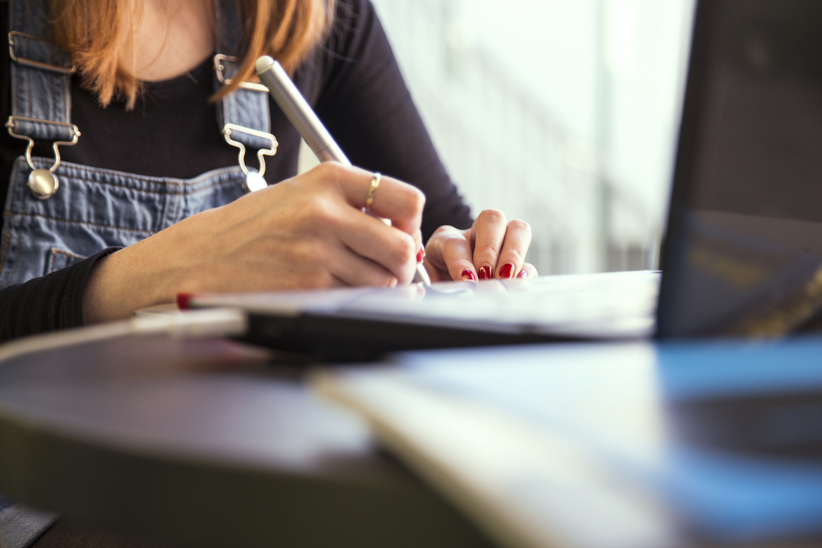 Das Bild zeigt den Oberkörper und die Hände einer an einem Tisch vor einem Laptop sitzenden Frau in Jeans-Latzhose, die etwas aufschreibt. 