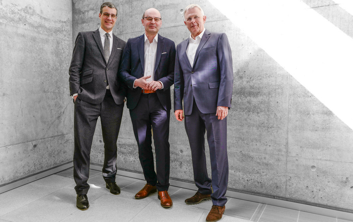 Drei Männer im Anzug stehen vor einer Betonwand an die ein Sonnenstrahl fällt