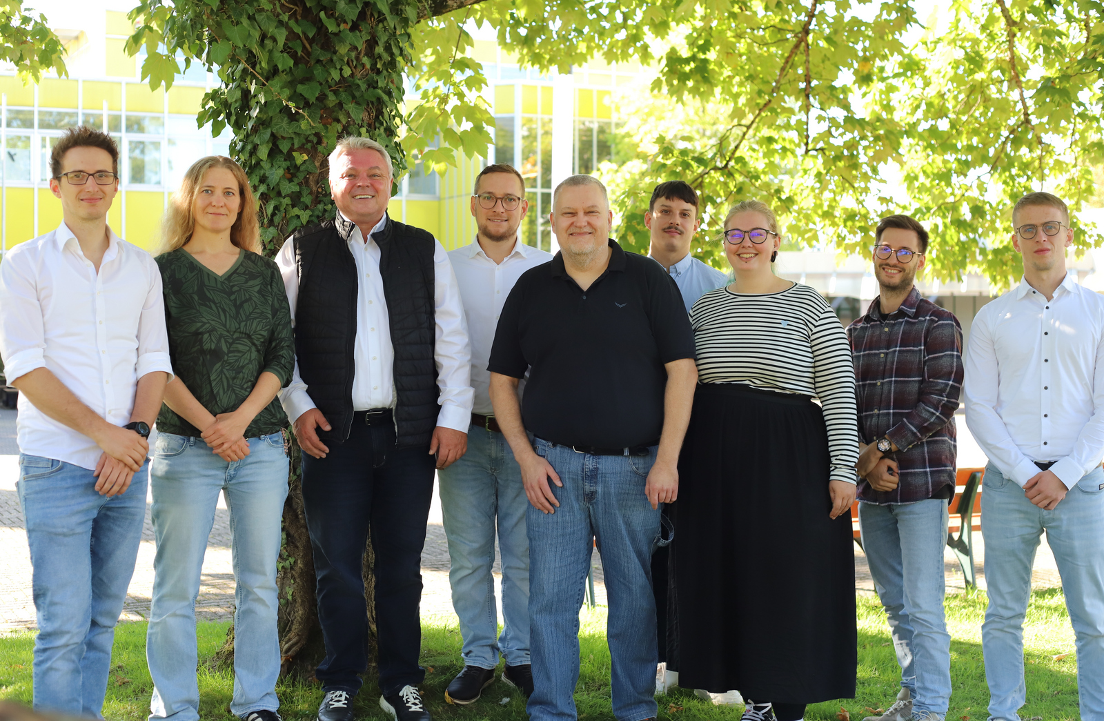 Gruppenfoto des Teams bei Sonnenschein unter einem Baum auf dem Rasen auf dem Campus