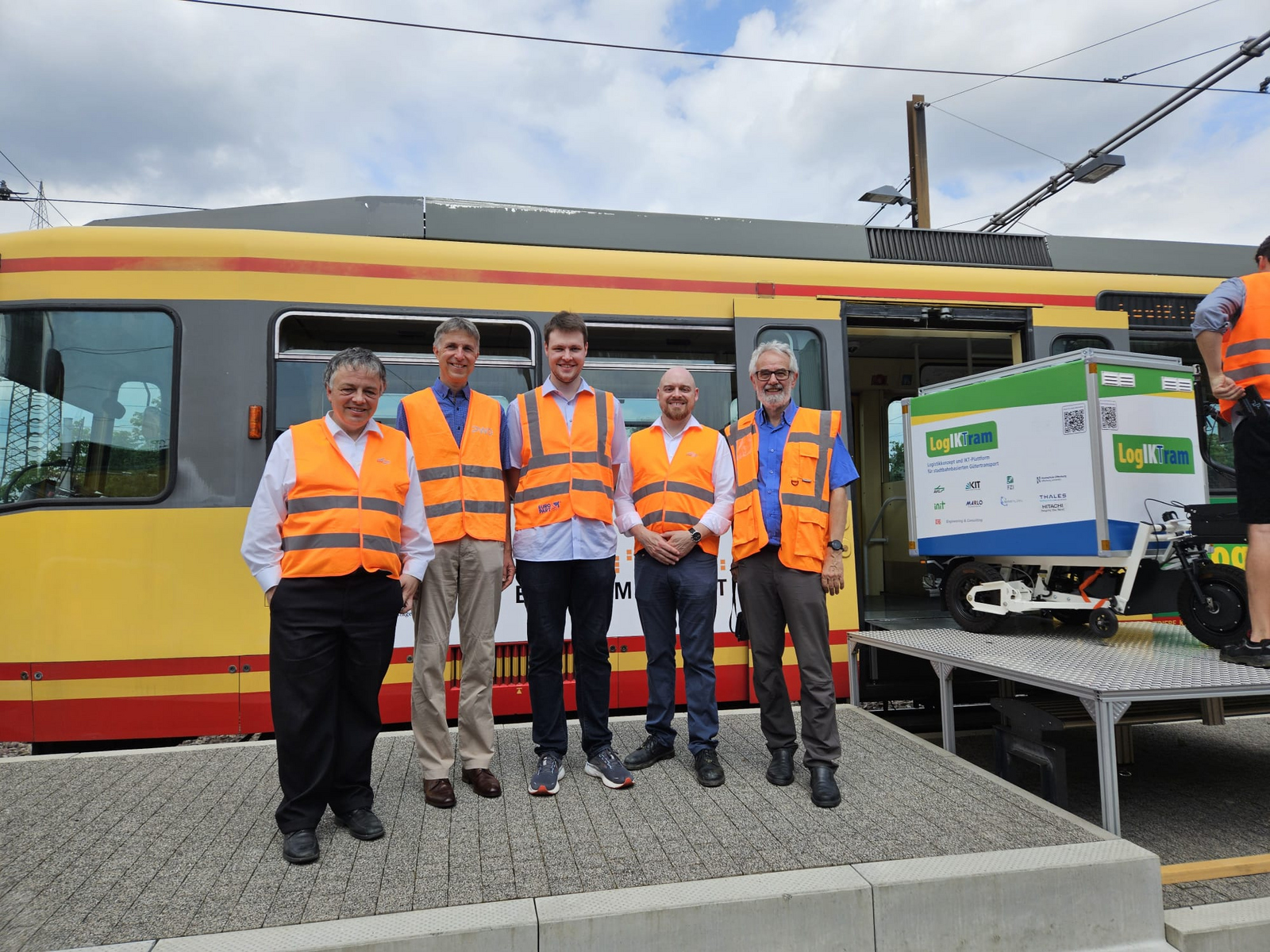 Fün Männer unterschiedlichen Alters in orangefarbenen Warnwesten stehen vor einem Straßenbahnwagen in den ein Lastenradanhänger hinfährt