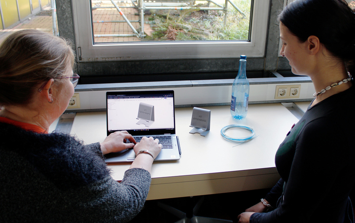 zwei junge Frauen sitzen an einem Laptop auf dem eine Konstruktionsskizze eines Smartphone-Halters zu sehen ist. Neben ihnen auf dem Tisch eine leere Plastikflasche, Filament und ein fertiger Smartphone-Halter aus dem 3D-Drucker