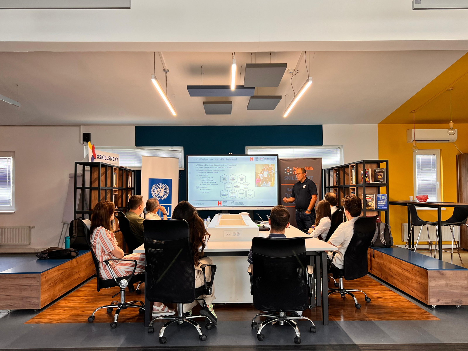 8 Personen an einem Tisch, mit Blickrictung zur einem großen Monitor neben dem eine Person präsentiert