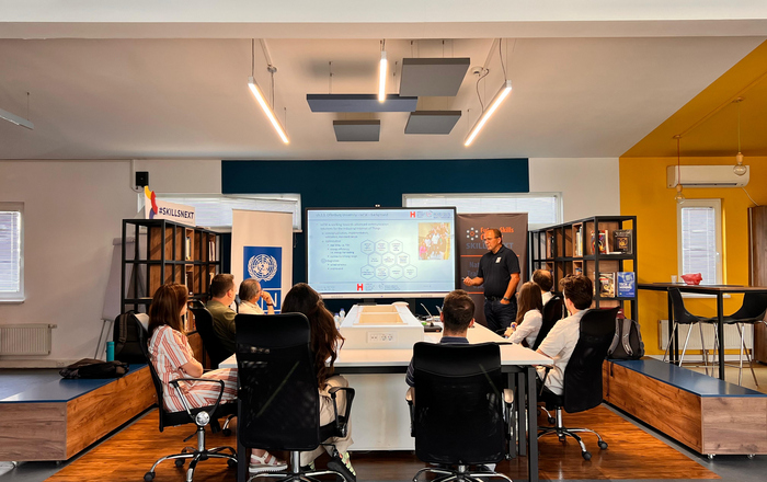 8 Personen an einem Tisch, mit Blickrictung zur einem großen Monitor neben dem eine Person präsentiert