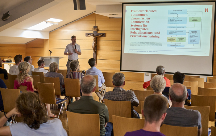 Mann steht neben Bildschirm vor sitzenden Zuhörenden