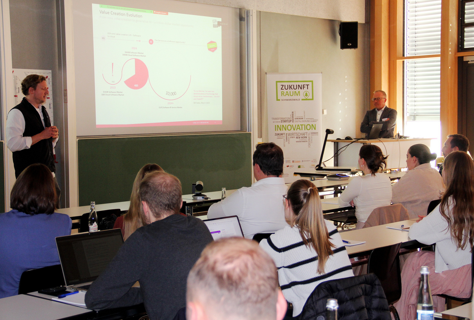 Zwei Männer stehen rechts und links einer Leinwand vor den sitzenden Zuhörenden und erläutern eine Präsentation auf der Leinwand