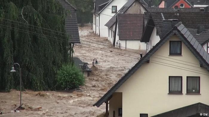 Eine braune Flutwelle bahnt sich ihren Weg zwischen Häusern und Bäumen hindurch 