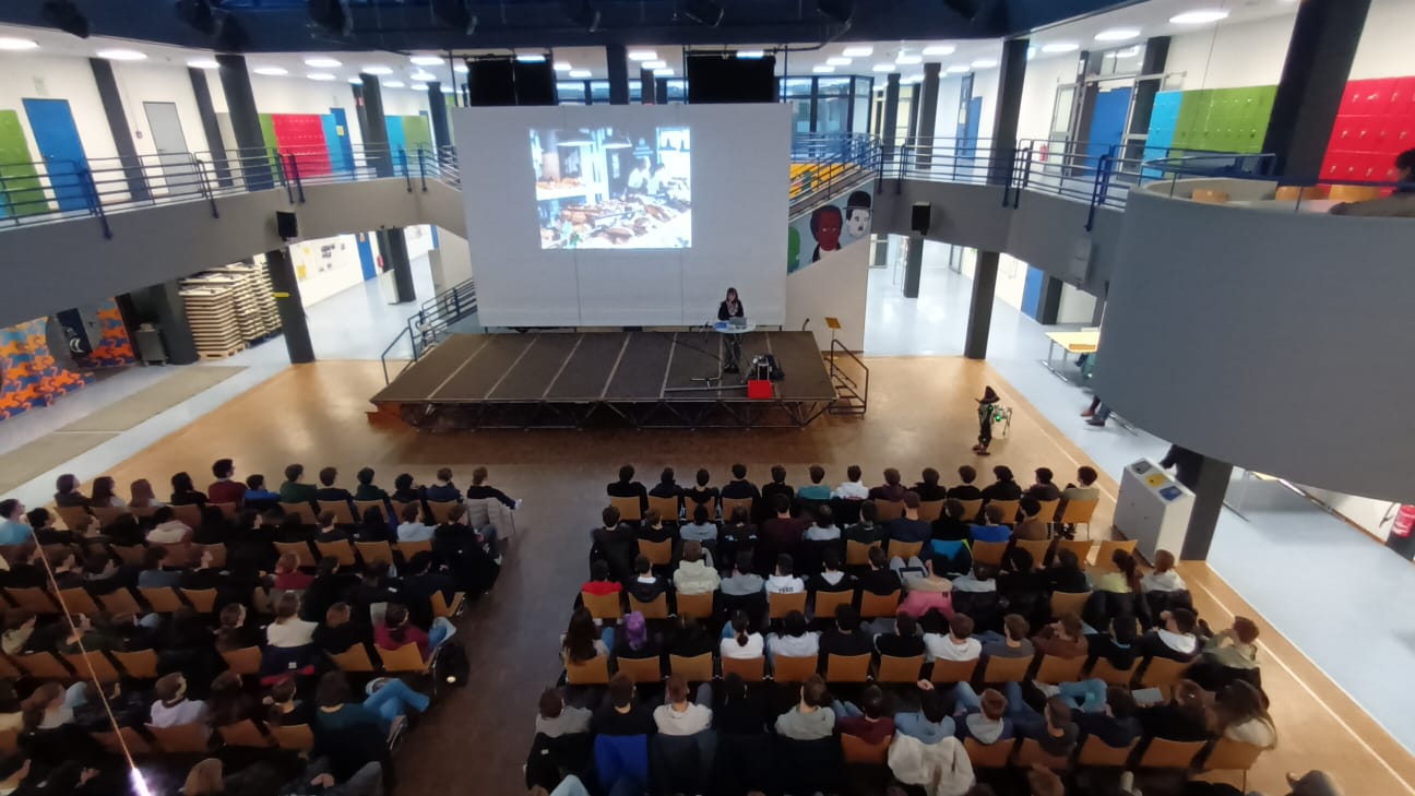 Ein Frau steht in einem großen Saal vor einer Leinwand und hält einen Vortrag vor sitzenden Zuhörenden