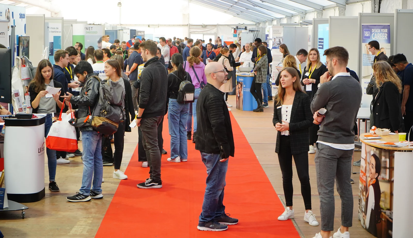 Blick ins Zelt der CAREER-Messe. Links und rechts die Messe-Stände der Unternehmen dazwischen flanieren zahlreiche Studierende, einige sprechen mit Unternehmenvertretern.